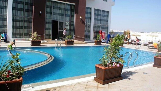Una piscina con un edificio moderno al fondo, una torre de salvavidas y sillas de descanso en la terraza.