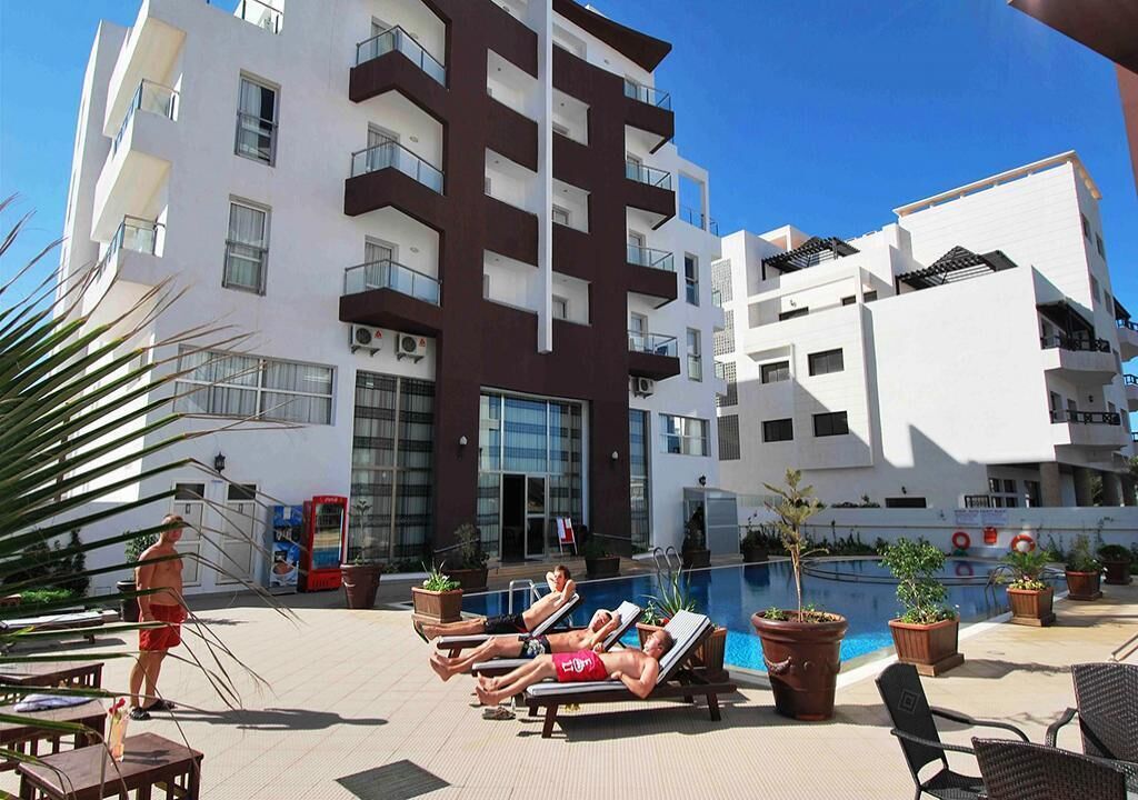 Un área junto a la piscina con sillones de descanso, una piscina y un edificio con balcones.