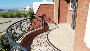 Un balcón exterior curvo con barandilla de metal decorativa, un muro de ladrillo y vistas a la playa y edificios a lo lejos.