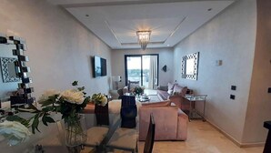 A modern living room with a glass dining table, a sofa, and a coffee table. There is a chandelier, a TV, and a large window with a view. - Residence Hivernage Founty 5165 (Agadir)