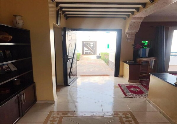 A hallway with a tiled floor leading to a door, a small rug, and a shelf with items on the left.