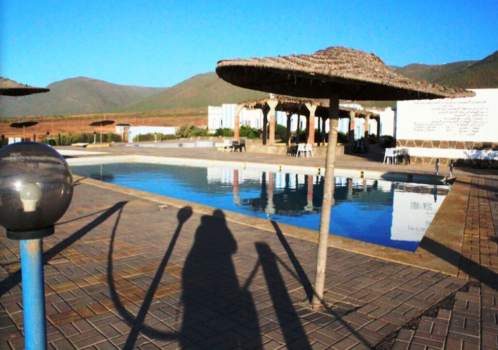 Un espace piscine agrémenté d’un parasol à toit de chaume, un ciel dégagé et des montagnes en arrière-plan.