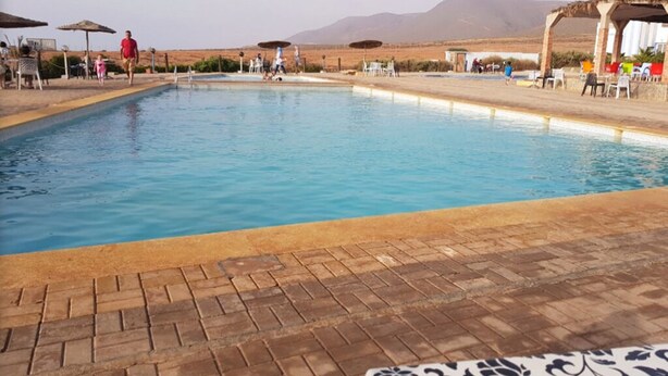 Ein Schwimmbecken mit Backstein-Terrasse, umgeben von Sonnenschirmen und Liegestühlen, vor einer Bergkulisse.