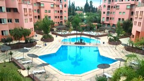 A swimming pool surrounded by lounge chairs and umbrellas, with a backdrop of pink buildings and greenery. - Residence Atlas Marrakech 5238 (Marrakech)