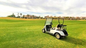 A golf cart on a well-maintained green field with a backdrop of residential buildings and palm trees. - Residence Marrakech Prestigia Golf City 5246 (Marrakech)