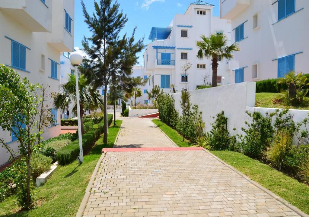 A pathway leading to a building with white walls and blue windows, surrounded by greenery and palm trees.