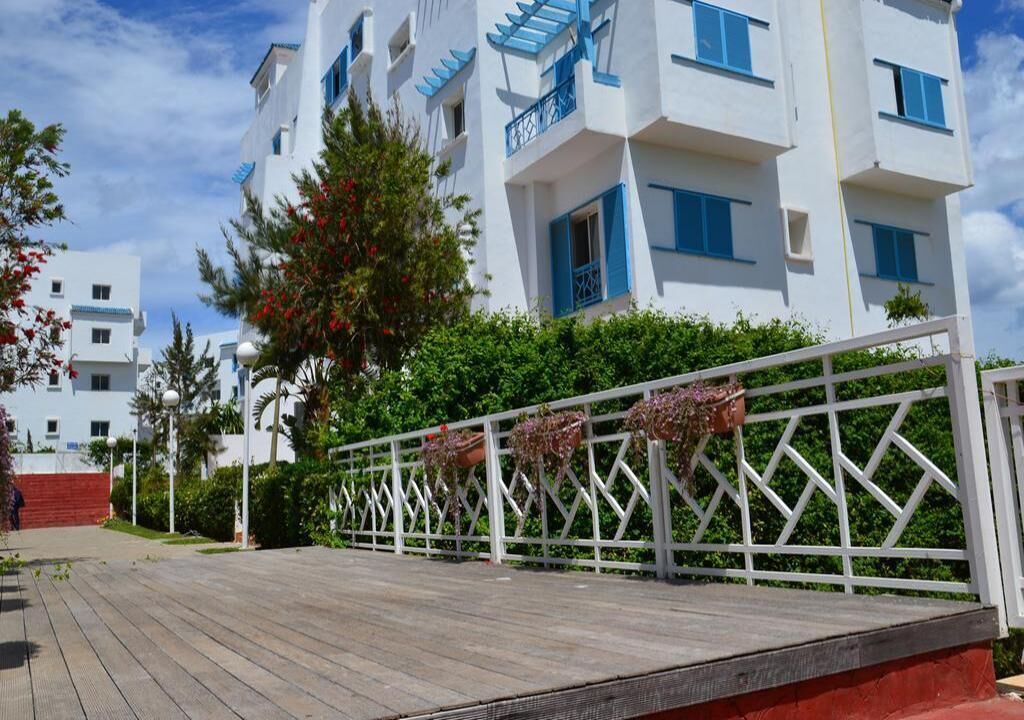 A modern building with white exterior and blue accents, a wooden deck, and a white fence with hanging flower pots.