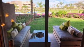 A modern living room with a glass partition, two sofas with cushions, a coffee table, and a view of a landscaped garden with palm trees and flowers. - Residence Marrakech Prestigia Golf City 5280 (Marrakech)