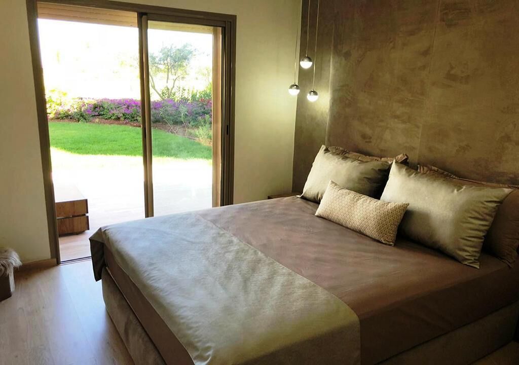 A bedroom with a large bed, a sliding glass door to a garden, and a wall-mounted light fixture.