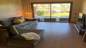A modern living room with a sofa, a flat-screen TV, and a sliding glass door leading to a balcony with outdoor furniture. - Residence Marrakech Prestigia Golf City 5280 (Marrakech)