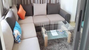 Een woonkamer met een beige hoekbank, een glazen salontafel en uitzicht buiten door een glazen deur.