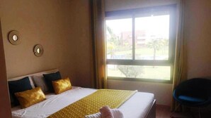 Una habitación de hotel con una cama grande, dos apliques, una ventana con vistas a edificios y zonas verdes, y una silla.