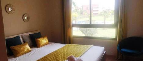 A hotel room with a large bed, two wall sconces, a window with a view of buildings and greenery, and a chair.