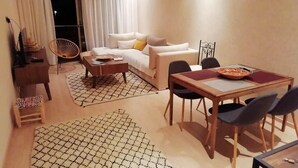 A modern living room with a sofa, coffee table, dining table, and chairs. There is a piano, a small stool, and a patterned rug. - Residence Marrakech Prestigia Golf City 5304 (Marrakech)