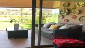 A modern living room with a sofa, cushions, a coffee table, and a patterned ottoman. A glass door leads to an outdoor area with a garden view.