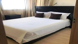 Ein ordentlich bezogenes Bett mit weißen Laken und zwei dunklen Kissen, ein Nachttisch aus Holz links daneben sowie ein Fenster mit durchsichtigen Vorhängen.