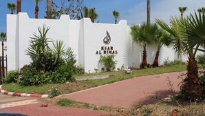 A white wall with the text "KSAR AL RIMAL" and a logo, surrounded by palm trees and greenery.