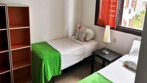 A small, tidy room with two single beds, a green blanket on one bed, a nightstand with a lamp, a window with red curtains, and a wooden wardrobe. - Residence Mangle 5332 (Cabo Negro)