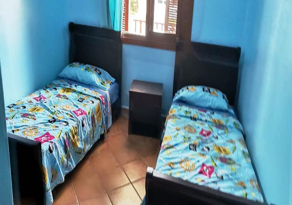 A room with two single beds, each with a blue bedspread featuring cartoon characters. The room has blue walls and a tiled floor.