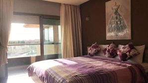 A hotel room with a large bed, a window with curtains, and a painting of a dress on the wall.