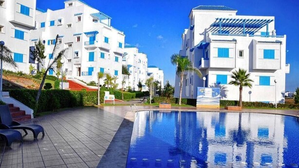 Zona de piscina con edificios blancos modernos y detalles azules, rodeada de palmeras y un cielo despejado.