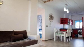 Una sala moderna con sofá, mesa de comedor y sillas. Se ve una cocina con armarios rojos y una lavadora al fondo.