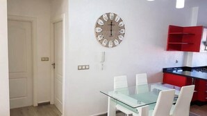 Un comedor con una mesa de vidrio y sillas blancas, un reloj de madera en la pared, y un armario de cocina rojo.