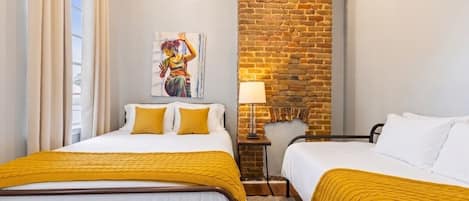 A hotel room with two beds, a brick accent wall, a painting, and a lamp.