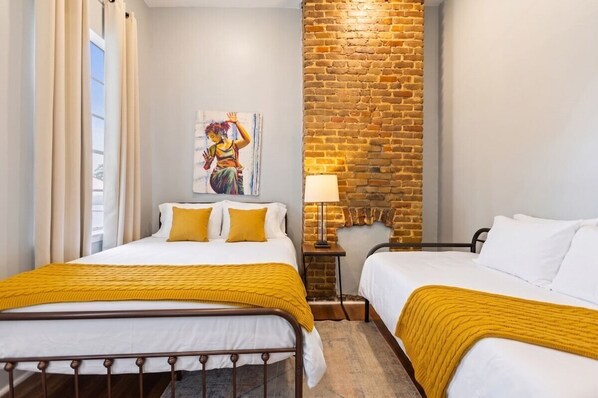 A hotel room with two beds, a brick accent wall, a painting, and a lamp.