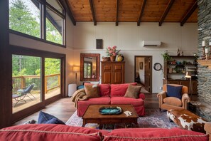 Una sala de estar acogedora con un sofá rojo, un sillón de cuero marrón y una mesa de centro de madera. Se ve una pared de piedra y una terraza de madera con barandilla.