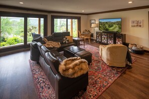 Un salón amplio con un sofá seccional grande, una mesa de centro y un televisor. Hay una ventana grande que ofrece vistas a la vegetación exterior.