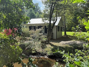 A small, single-story building with a metal roof, surrounded by lush greenery and a small stream.