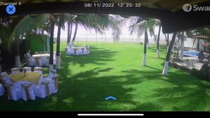 A covered outdoor area with tables and chairs, palm trees, and a view of the ocean.