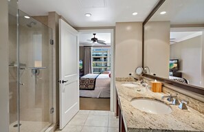 A hotel bathroom with a glass shower, a large mirror, a granite countertop, and a bed visible through an open door.