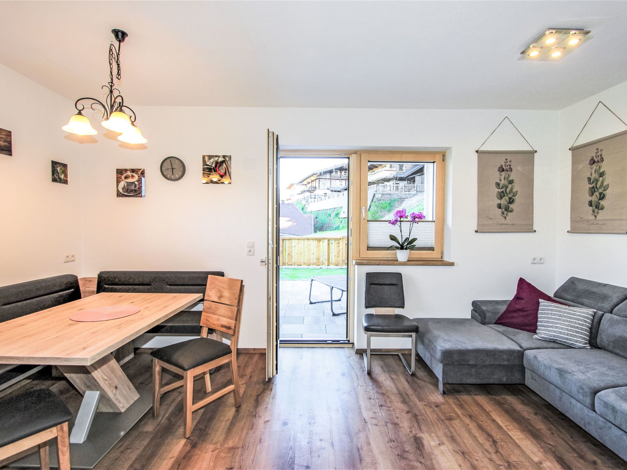 A modern living room with a wooden dining table, a grey sofa, and a bench. There is a window with a view of a garden and a balcony.