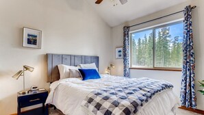 Une chambre à coucher comprenant un lit, des tables de chevet, une commode, un ventilateur de plafond, une fenêtre avec des rideaux et un tableau encadré accroché au mur.