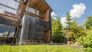 A multi-story wooden building with large windows, a balcony, and a garden area with flowers and trees. - Vacation Home "Haus der Sinne Bregenzerwald" with Mountain View & Balcony (Egg-Kaltenbrunnen)