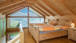 A wooden bedroom with a large window, a bed, and a nightstand. - Vacation Home "Haus der Sinne Bregenzerwald" with Mountain View & Balcony (Egg-Kaltenbrunnen)