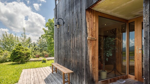 A wooden building with a wooden deck, a bench, and a glass door with a sign. - Vacation Home "Haus der Sinne Bregenzerwald" with Mountain View & Balcony (Egg-Kaltenbrunnen)
