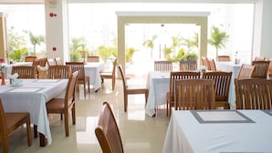 Un restaurante con varias mesas y sillas, amplias ventanas que ofrecen vista a palmeras y un interior luminoso.