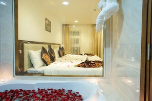 A hotel room with a large bathtub filled with rose petals, a bed with decorative pillows, and a view of the city through a glass door. - Centre Hotel (Da Nang Municipality)