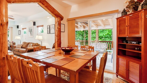 A spacious dining area with a wooden table and chairs, a large window with a view of greenery, and a wooden cabinet with shelves.