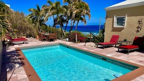 A rectangular swimming pool with a tiled edge, surrounded by a patio area with red lounge chairs and a table. - Stunning Paradise Overlook - Incredible Views from Every Room (Christiansted)