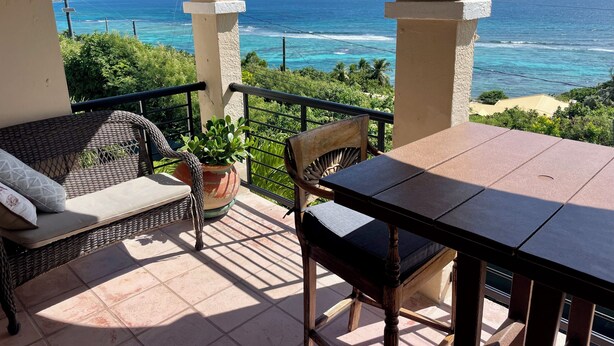 A balcony with a wicker sofa, a wooden chair, and a table, overlooking a turquoise sea and greenery. - Stunning Paradise Overlook - Incredible Views from Every Room (Christiansted)