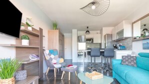 A modern living room with a sofa, a coffee table, and a TV. There is a kitchen in the background with bar stools.