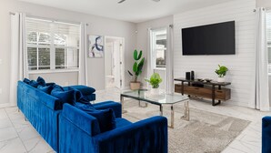 A modern living room with a blue sofa, a glass coffee table, a ceiling fan, and a flat-screen TV mounted on the wall. - Themed Games, Theater Room | Screened Pool Area (Davenport)