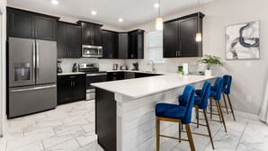 A modern kitchen with dark cabinets, stainless steel appliances, a white island with bar stools, and a large window with blinds. - Themed Games, Theater Room | Screened Pool Area (Davenport)