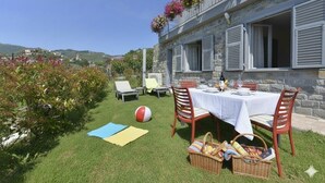 A stone building with a balcony adorned with flowers, a dining area with a table set for two, and a grassy lawn with a beach ball and picnic setup. - Residenza Cantalupo – Relax Oasis with PoolAp37 (Imperia)