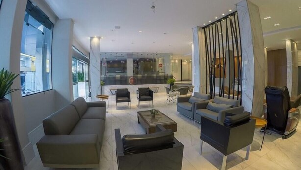 Una sala de estar moderna con un sofá, sillones y una mesa de centro. Se ve un ventanal grande a la izquierda y una cocina al fondo.