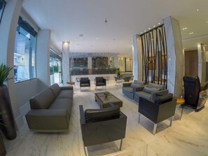 Un salon moderne avec un canapé, des fauteuils et une table basse. Une grande fenêtre se trouve à gauche, et on aperçoit un coin cuisine au fond.